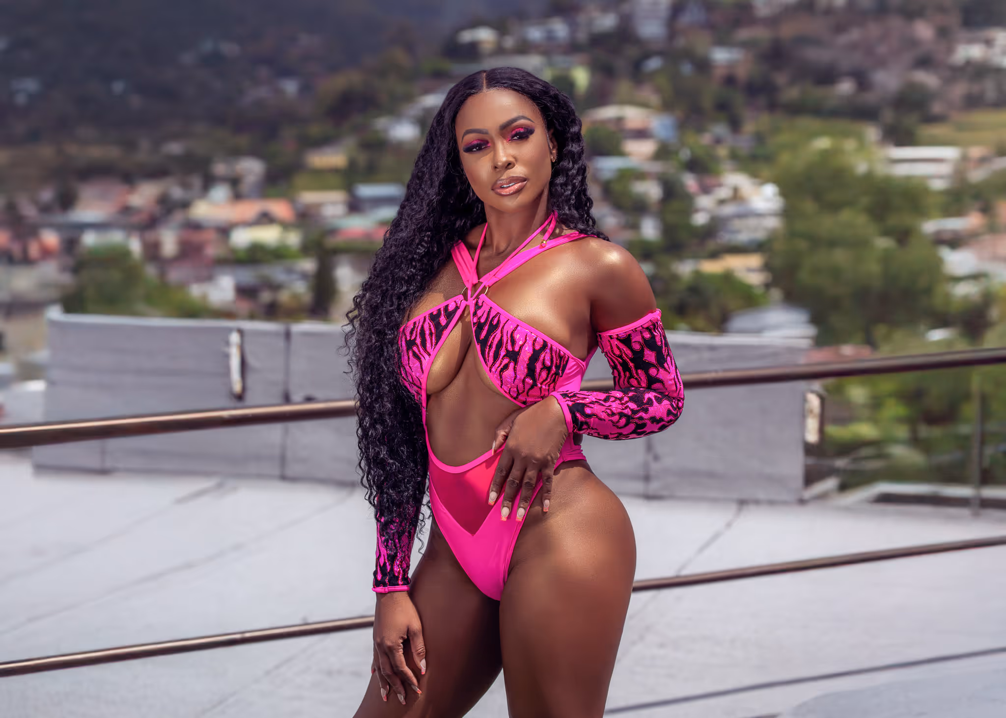 Woman wearing a vibrant pink cut-out swimsuit with flame patterns posing outdoors on a rooftop with a blurred town and hills in the background.