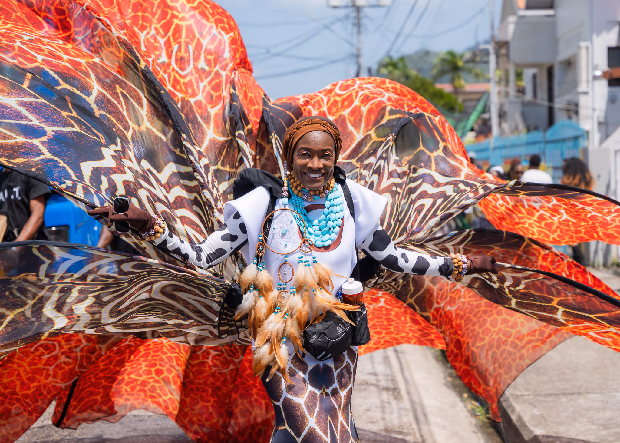 Person smiling in a colorful giraffe-patterned carnival costume with large orange and black wings, wearing turquoise beads and holding a dreamcatcher.