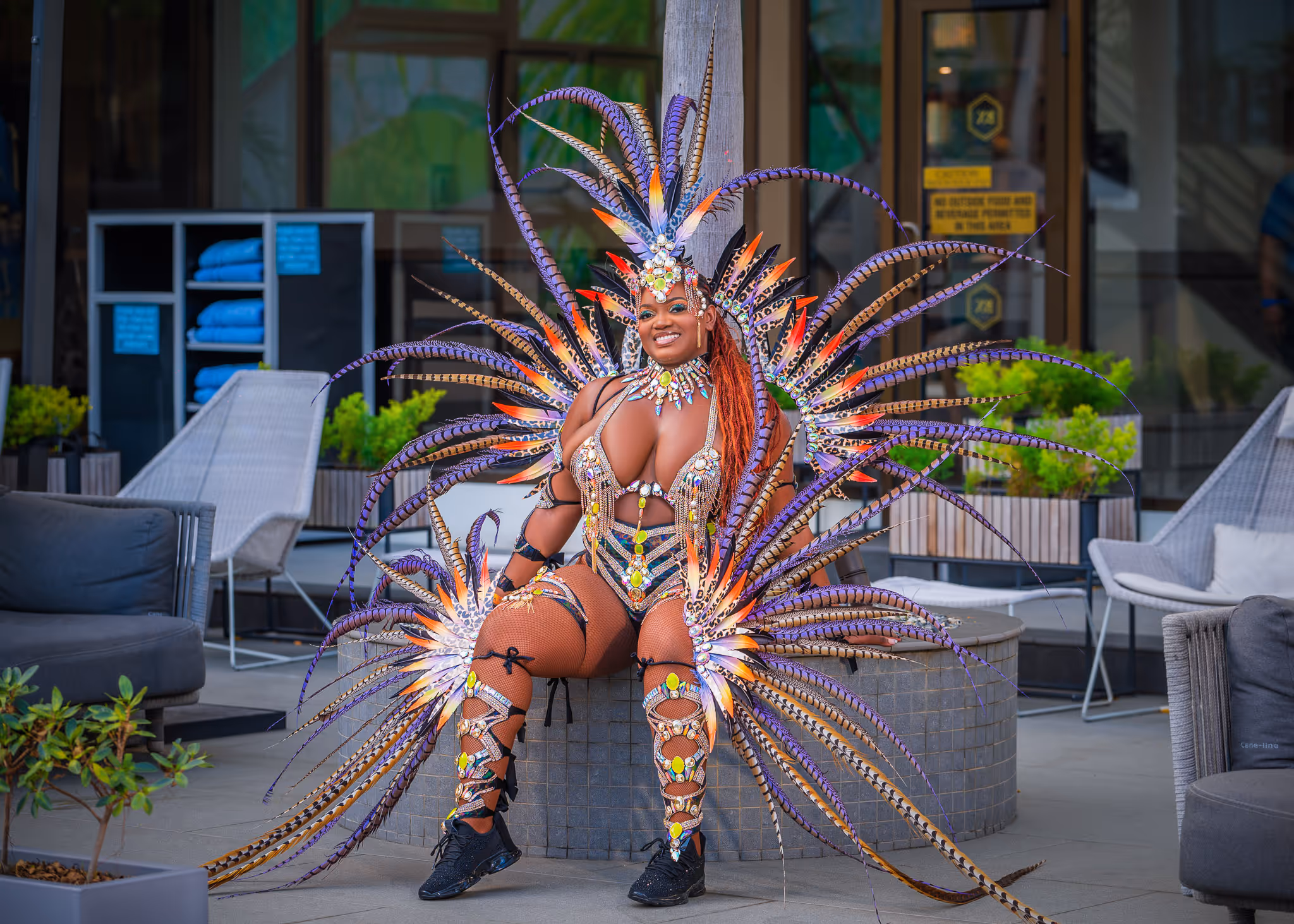 Smiling woman in an elaborate carnival costume with colorful feathers and gemstones sitting outdoors.