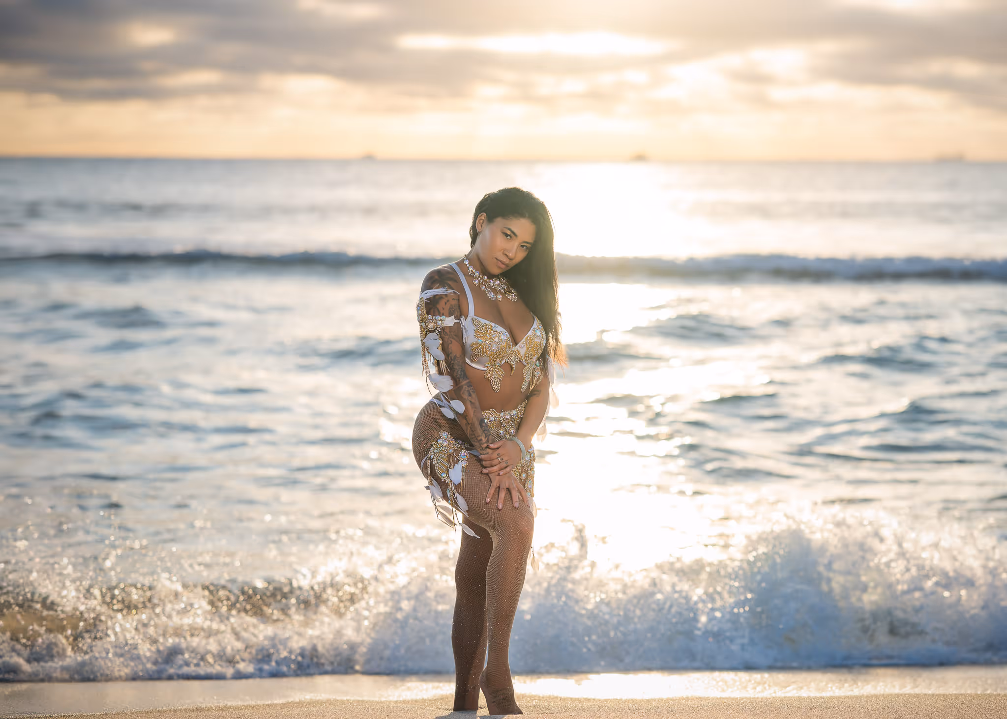 Woman wearing decorative carnival costume standing on a beach at sunset with ocean waves behind her.