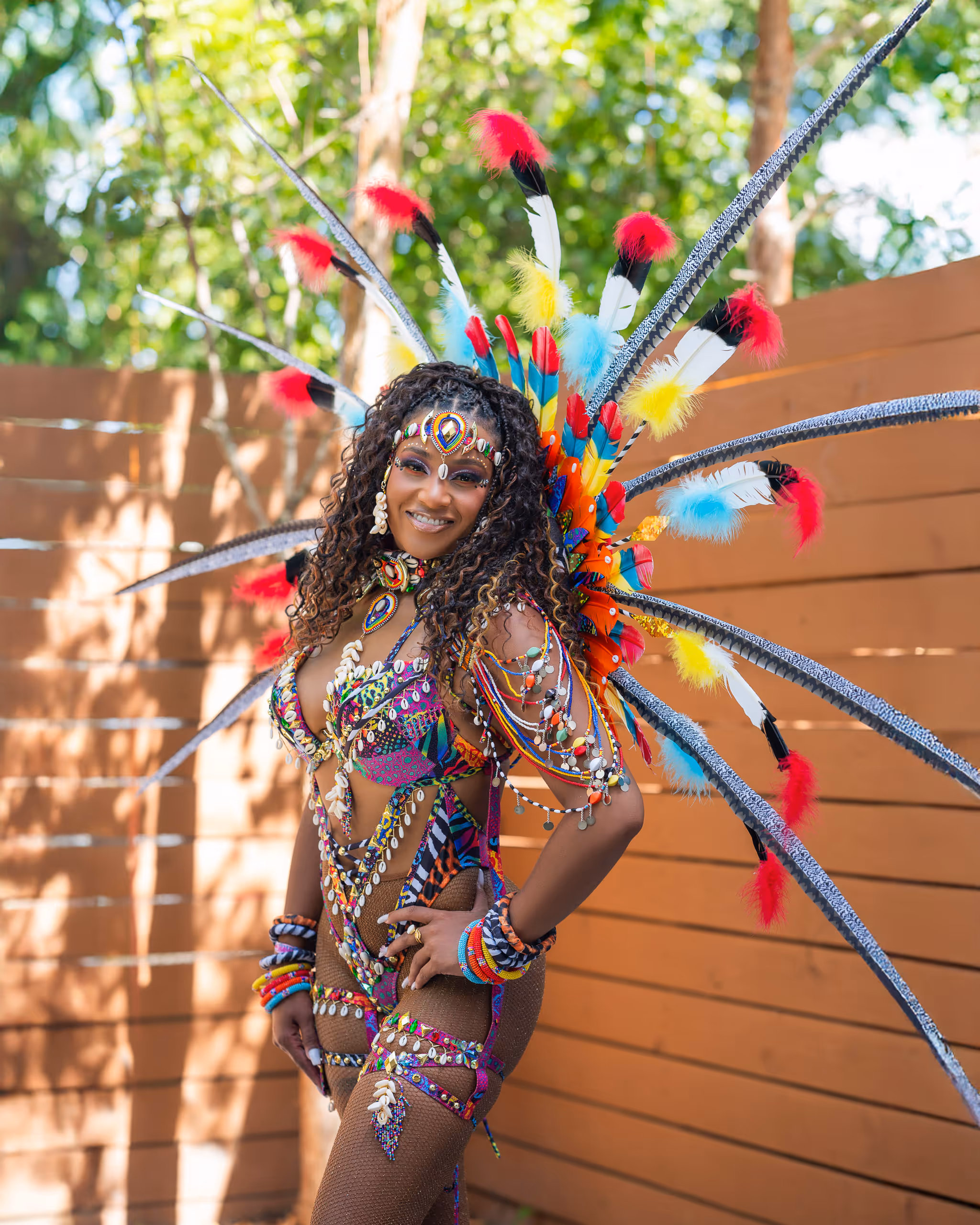 Smiling woman wearing a colorful carnival costume with feathers, beads, and shells standing outdoors.