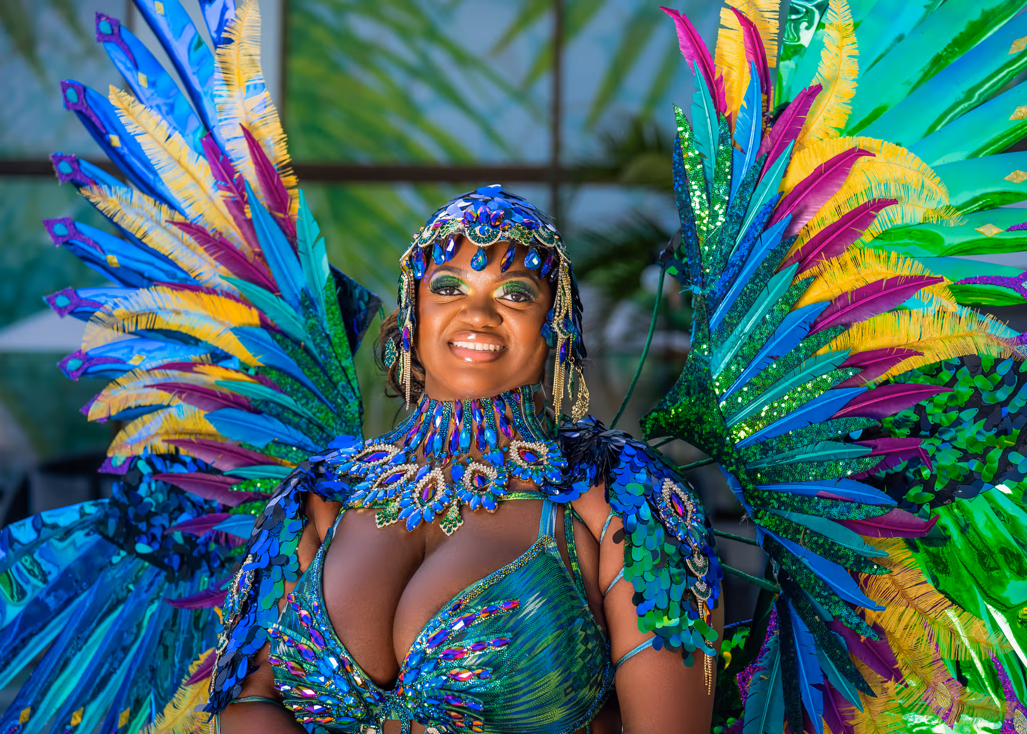 Smiling woman wearing an elaborate blue, green, yellow, and purple feathered carnival costume with sequins and jewels.