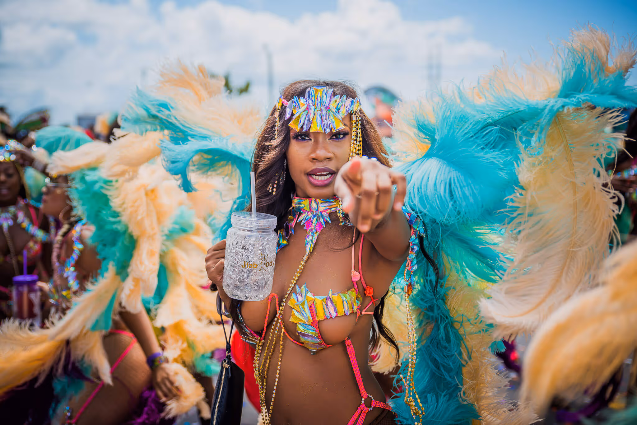 Woman in colorful carnival costume with large feathered wings holding a clear cup and pointing at the camera.