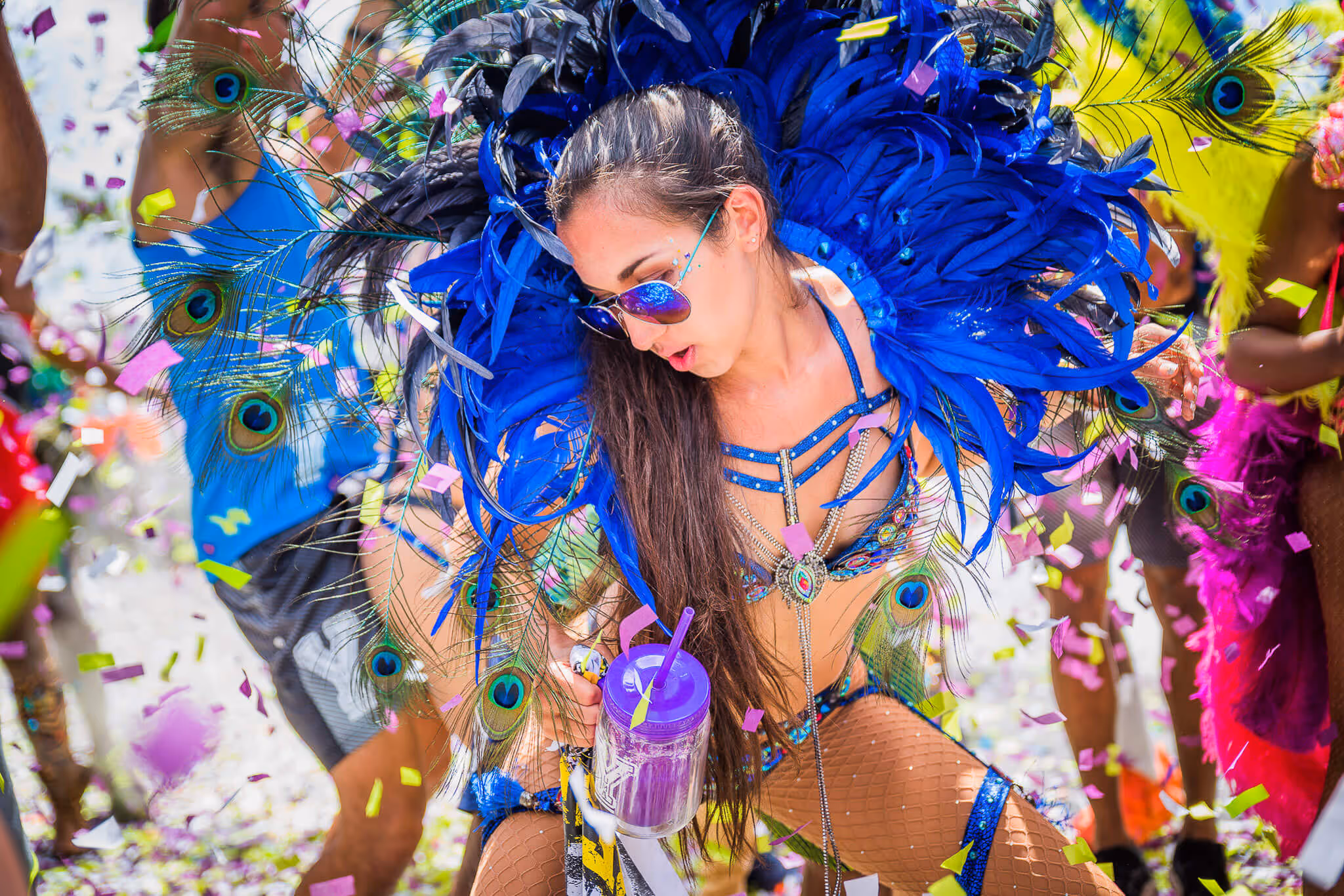 Woman wearing vibrant blue feathered costume with peacock feathers dancing at a colorful carnival with confetti falling around.