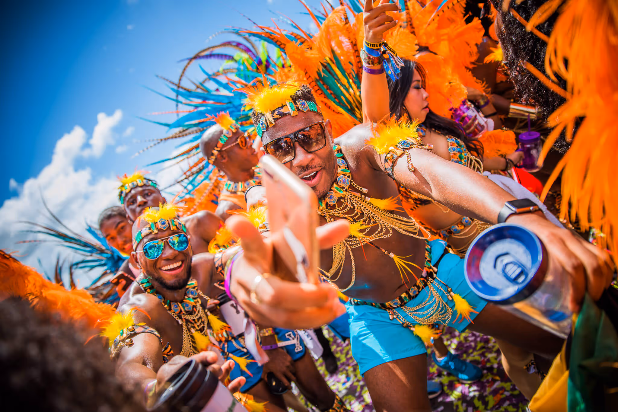 Group of people dancing energetically in colorful carnival costumes with orange feathers and blue accessories under a bright blue sky.