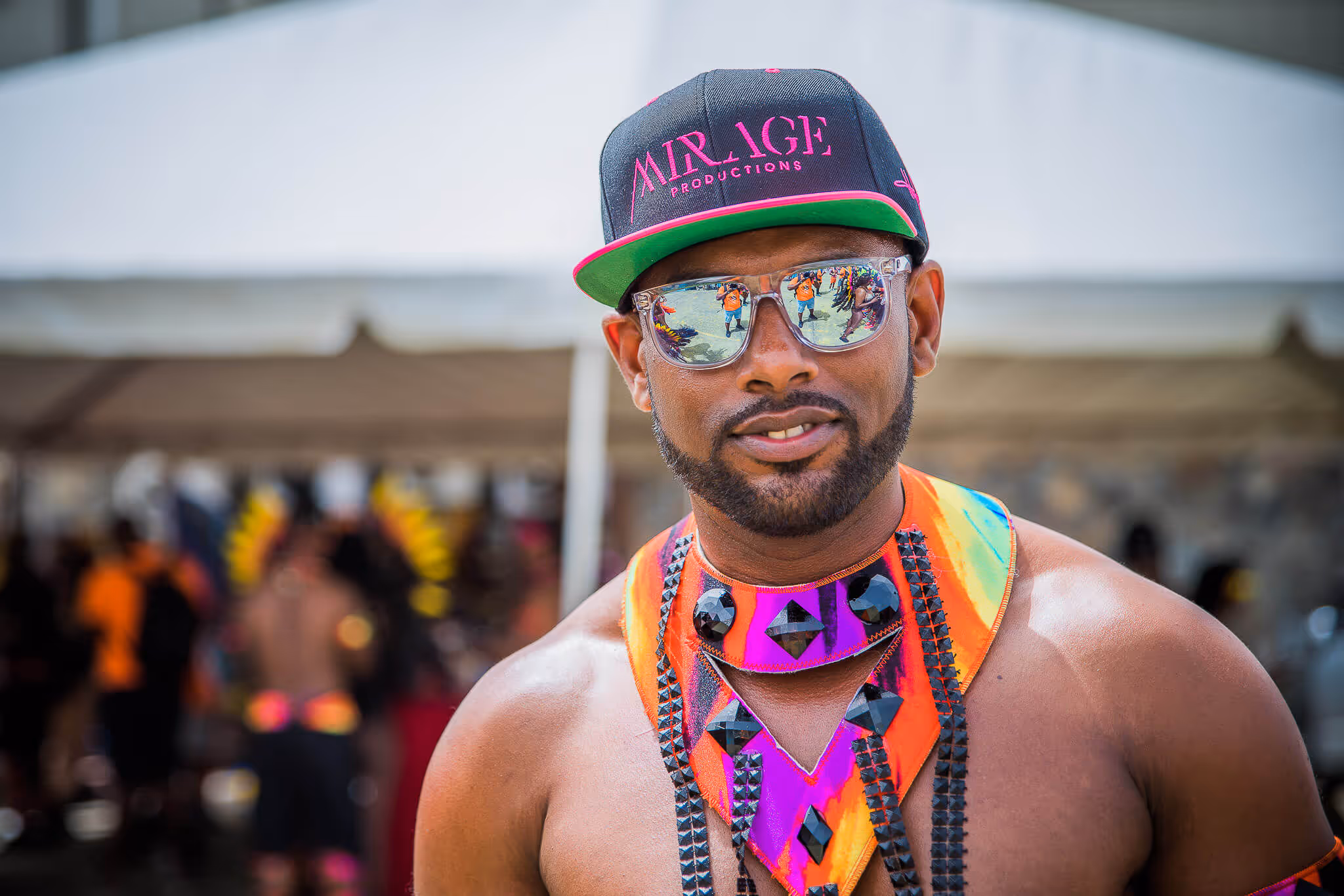 Man wearing a black and pink Mirage Productions cap, reflective sunglasses, and a colorful carnival costume with black bead embellishments.