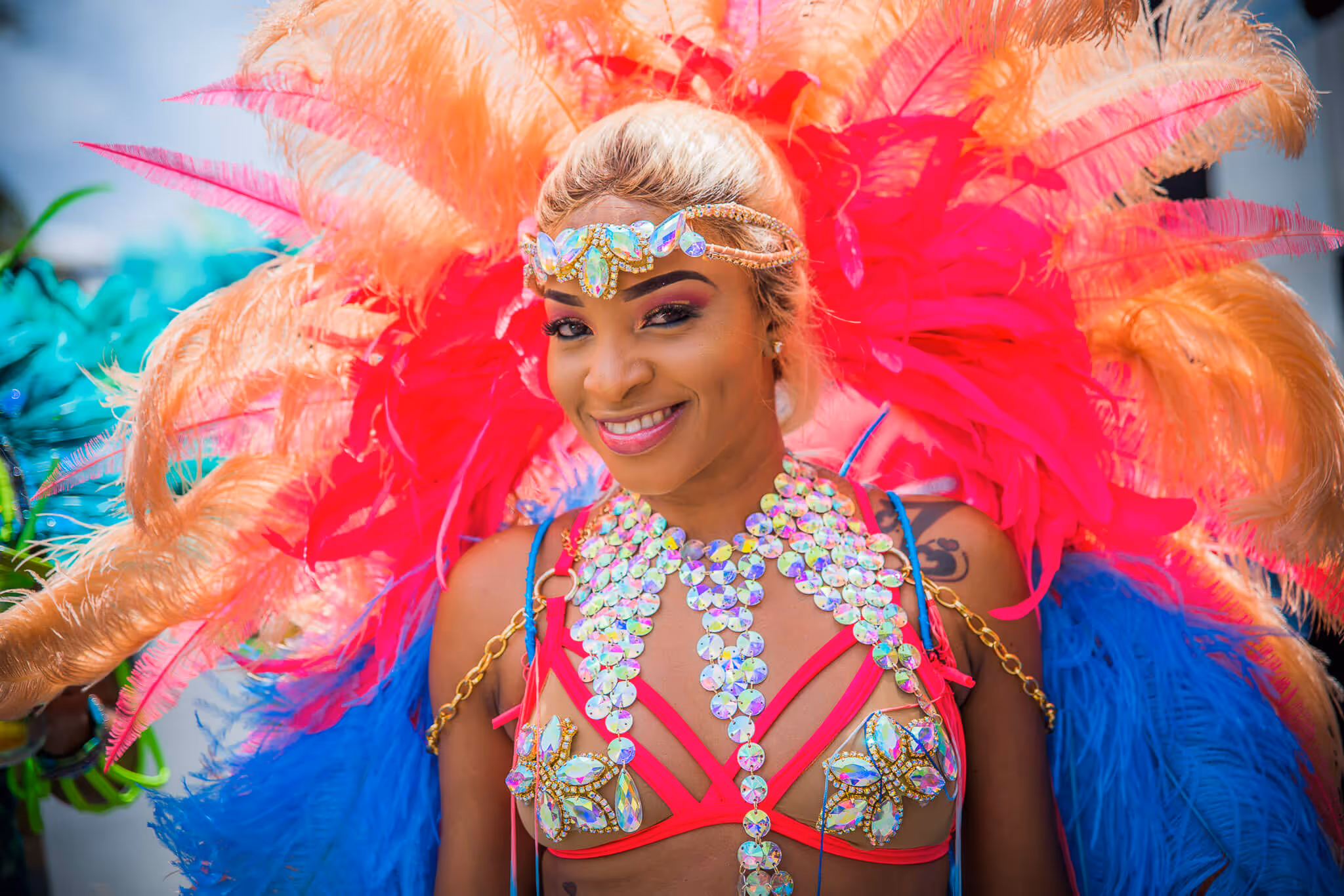 Smiling woman wearing a vibrant carnival costume with bright pink, orange, and blue feathers and sparkling decorative jewelry.
