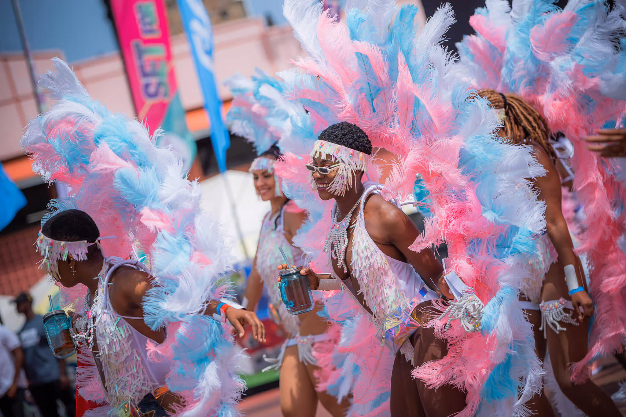 People dancing in colorful carnival costumes adorned with pink, blue, and white feathers and beads, holding drinks during a street parade.