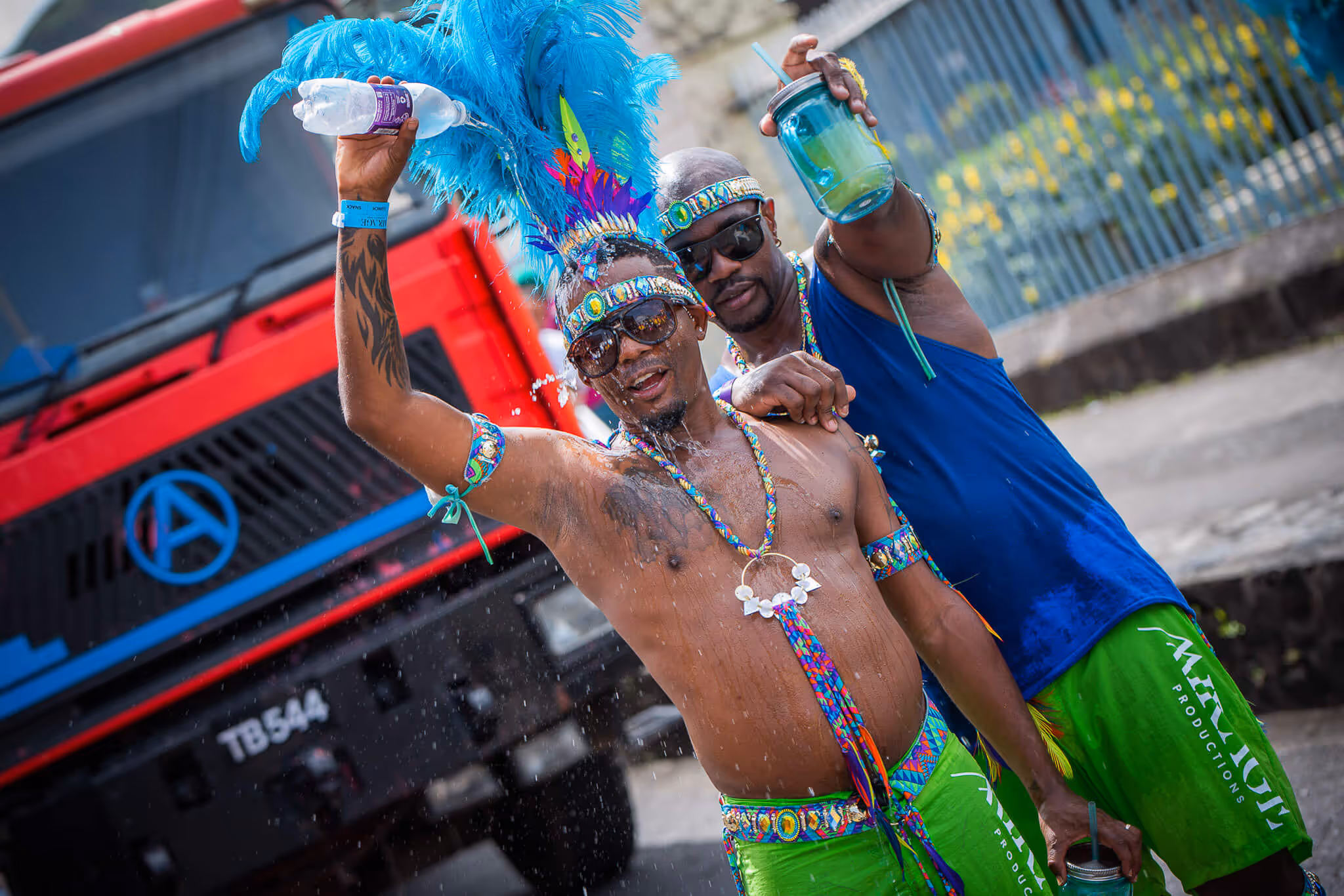 Two men dressed in colorful carnival attire with blue feathers and green shorts celebrating and holding drinks near a red truck.