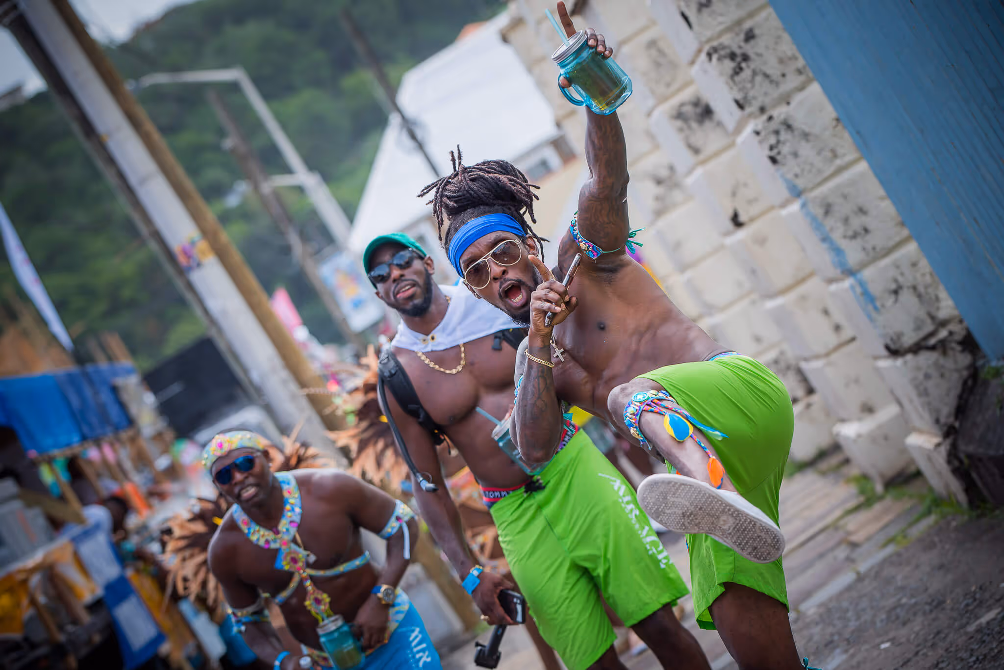 Three men in colorful carnival attire dancing and holding drinks on a street with stone walls.