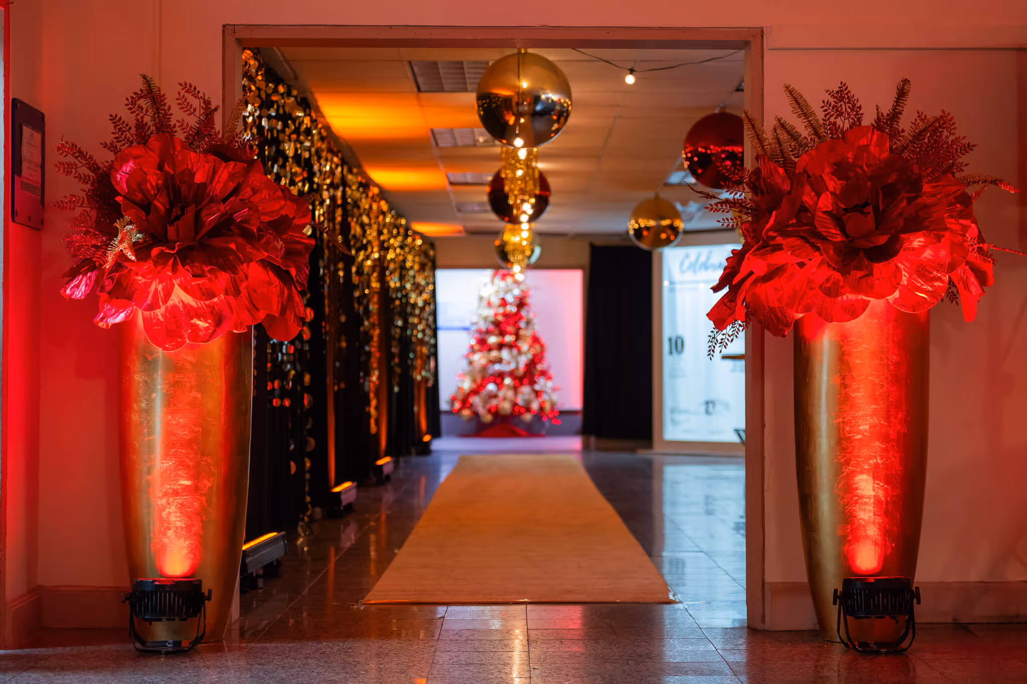 Indoor hallway decorated with large red floral arrangements in tall gold vases, illuminated by red uplighting, leading to a Christmas tree at the end.