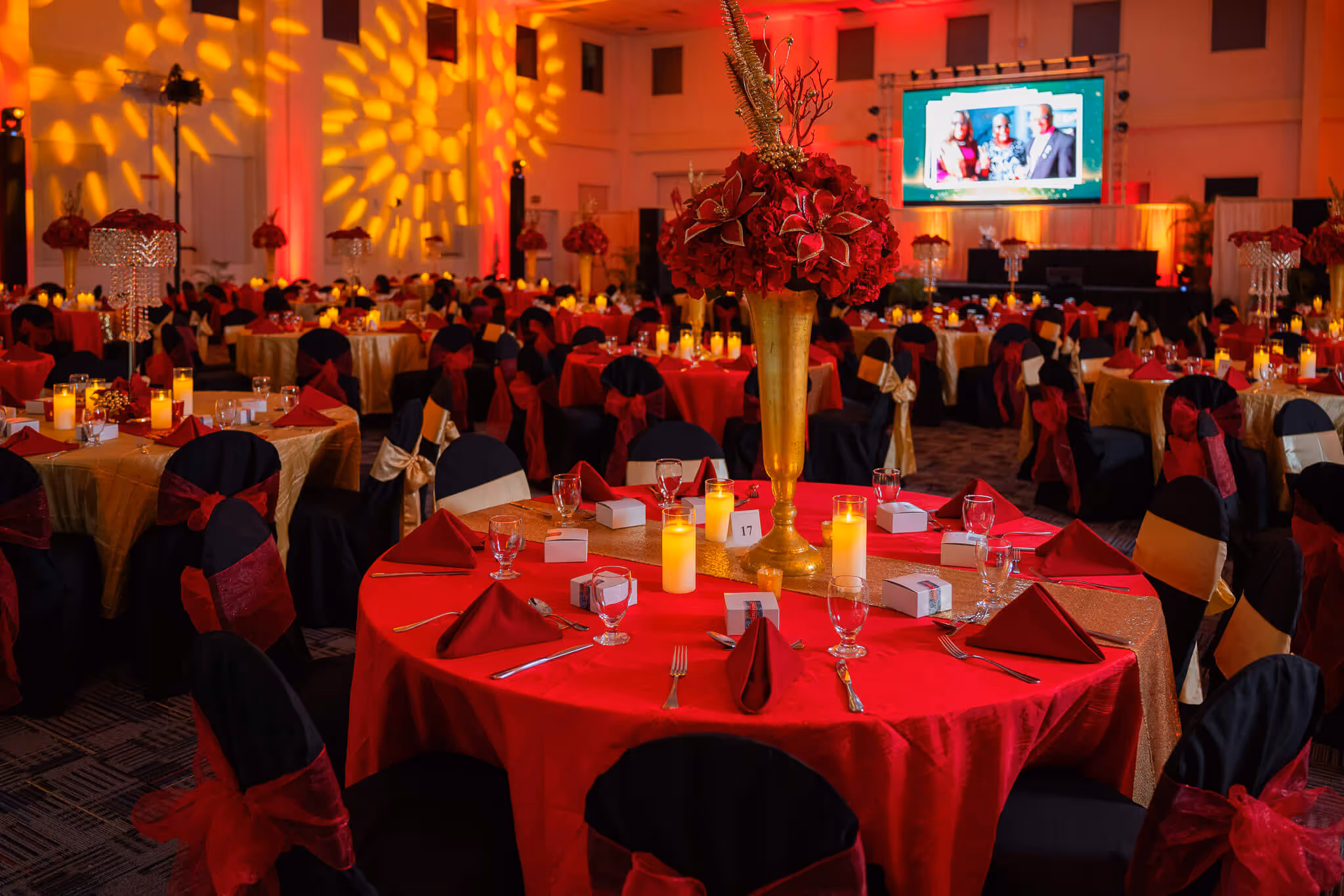 Large banquet hall decorated with red and gold table settings, black chairs with red and gold bows, tall floral centerpieces, and warm candle lighting.