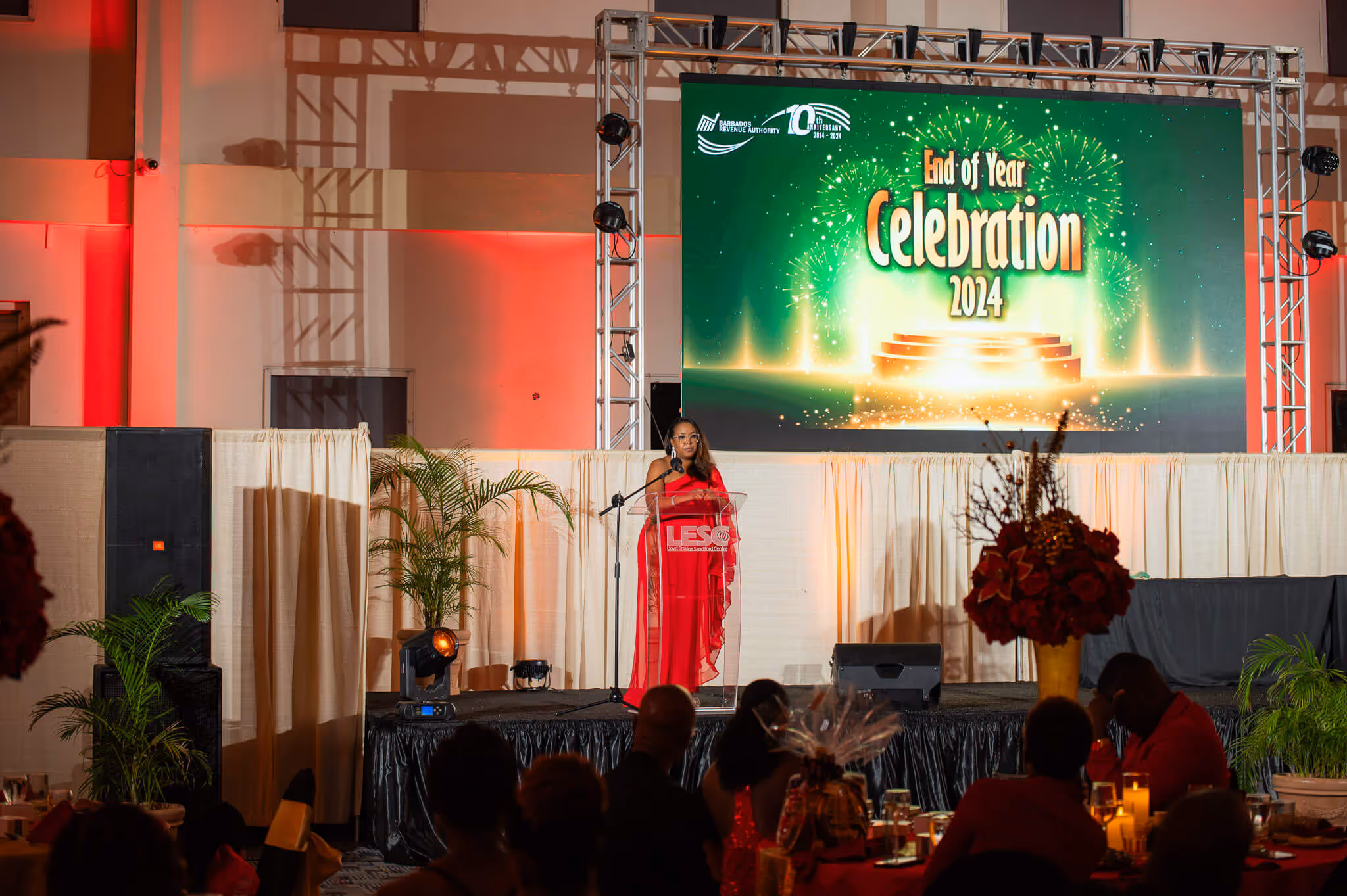 Woman in red dress speaking at a podium on a stage with a large screen displaying 'End of Year Celebration 2024' in the background.