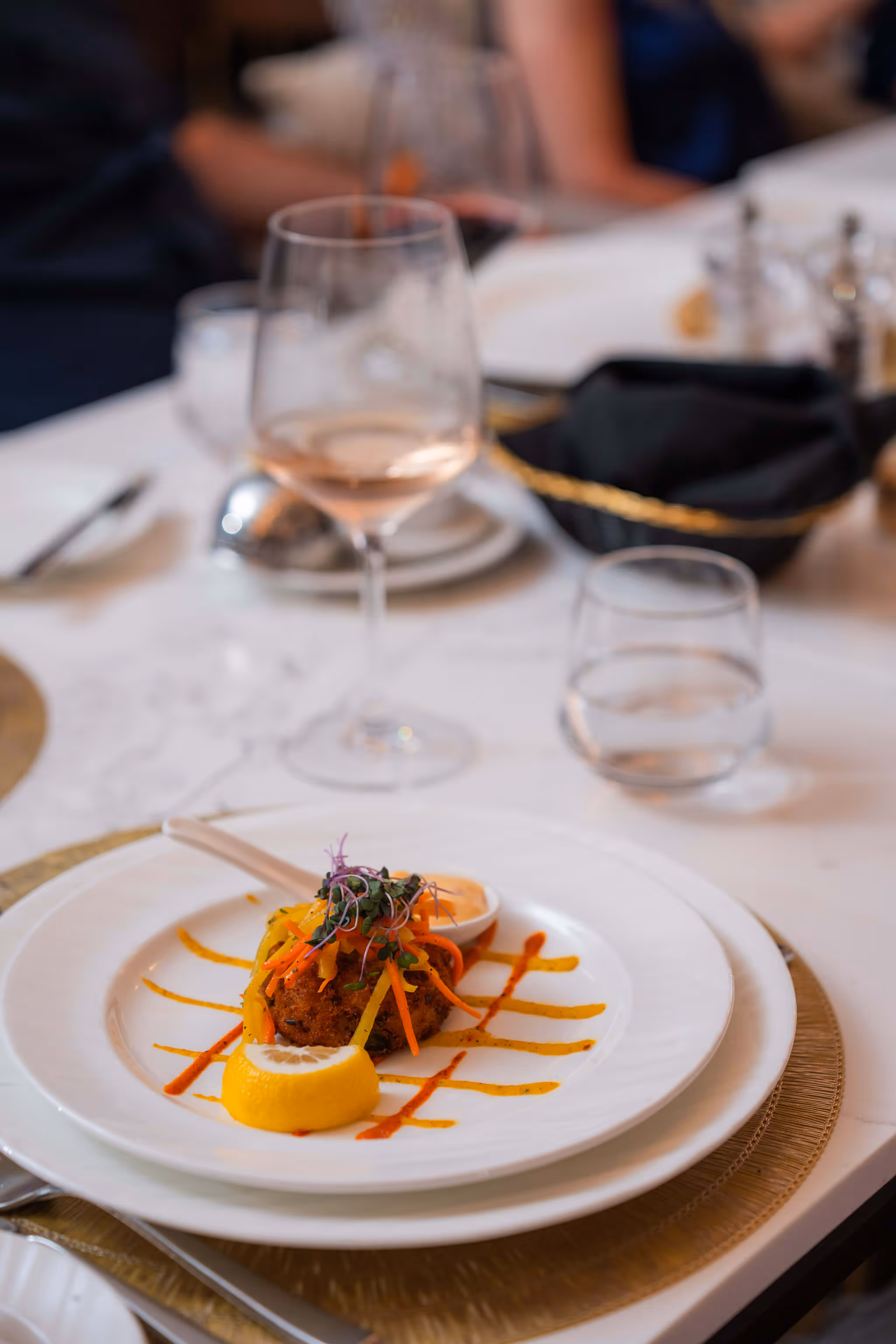 Gourmet plated dish with a crab cake topped with julienned vegetables and microgreens, served with a lemon wedge and sauce on a white plate.