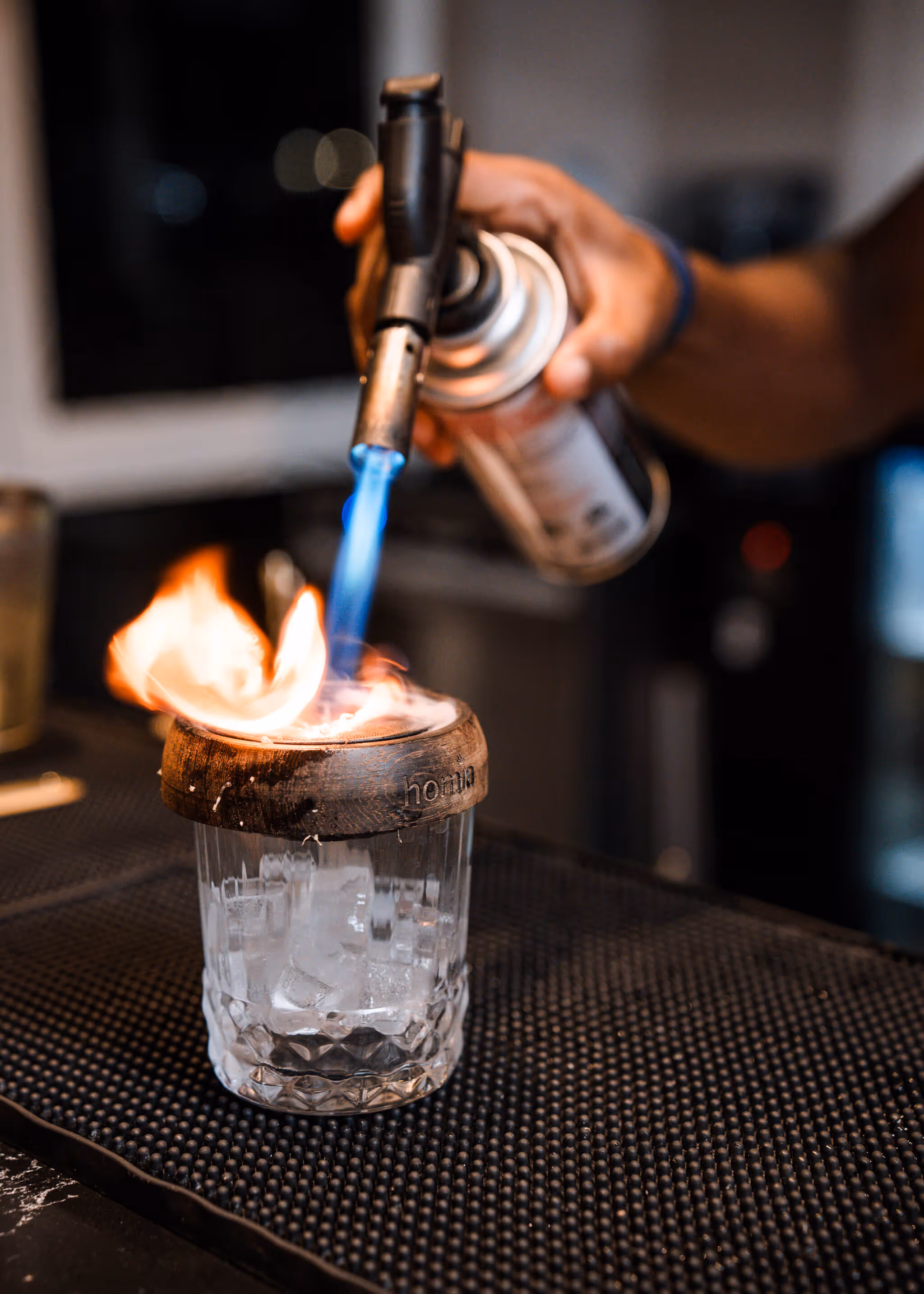 A hand uses a blowtorch to ignite the rim of a glass filled with ice on a bar mat.