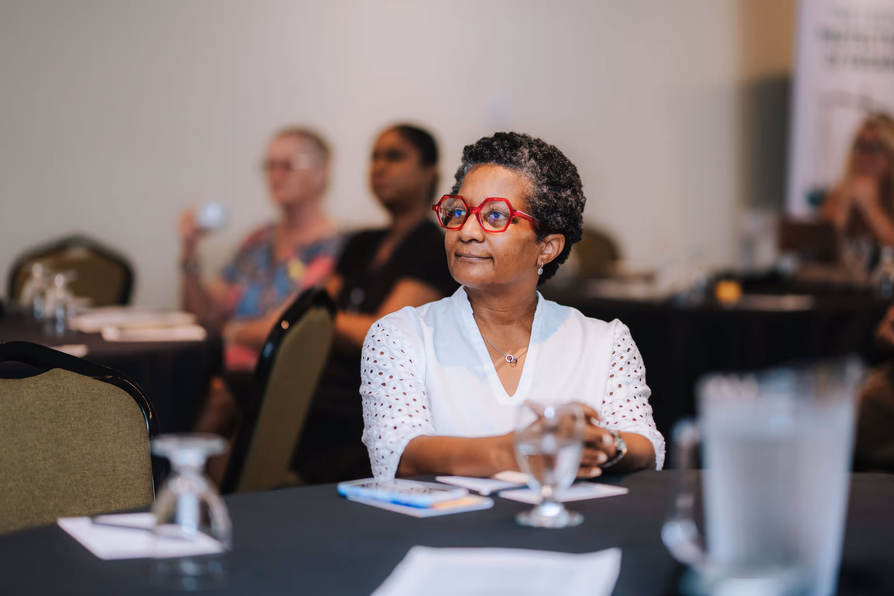 Woman with short curly hair and red glasses sitting at a conference table, attentively listening.