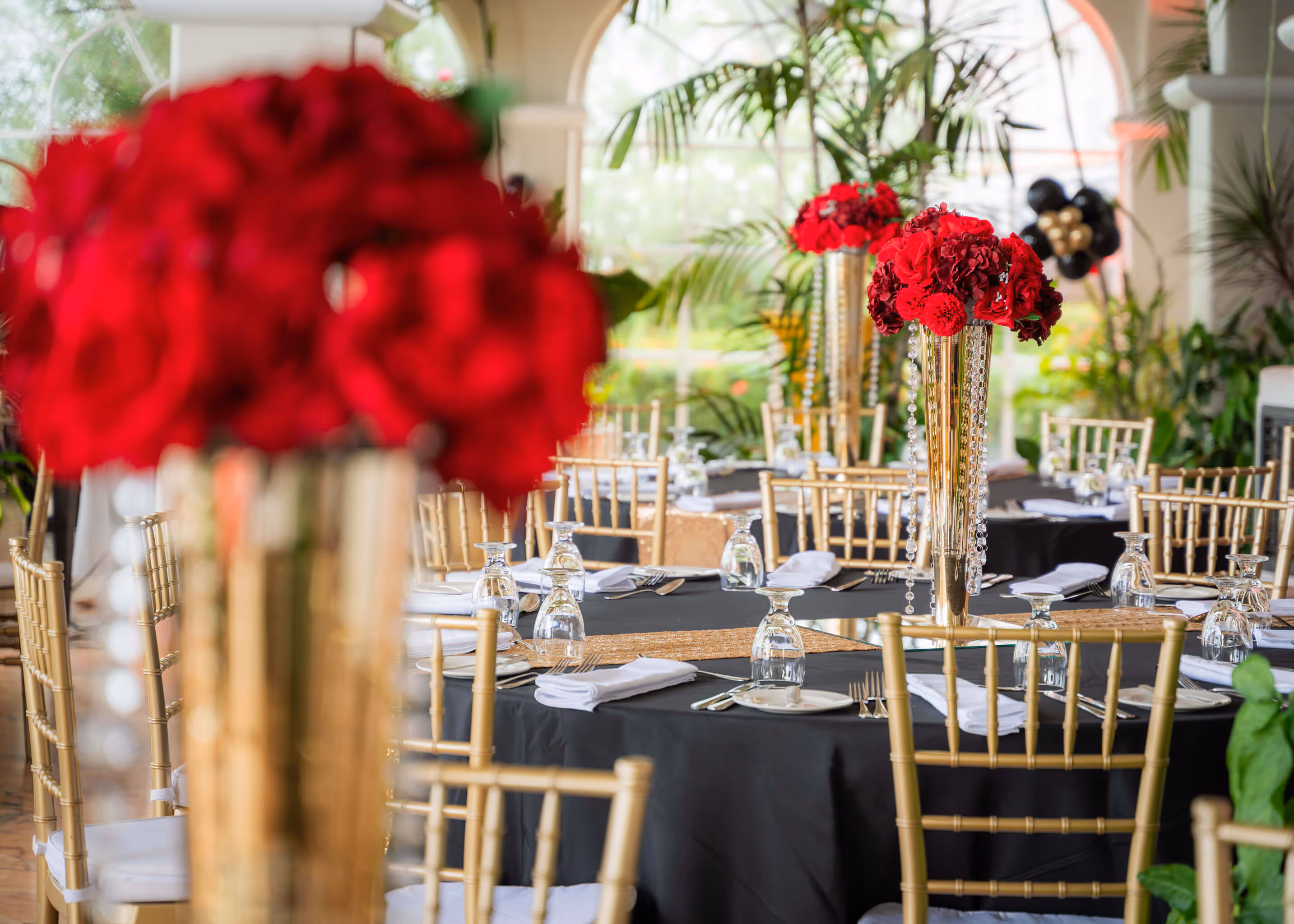 Elegantly set dining tables with black tablecloths, gold chairs, and tall gold vases holding red flower centerpieces.
