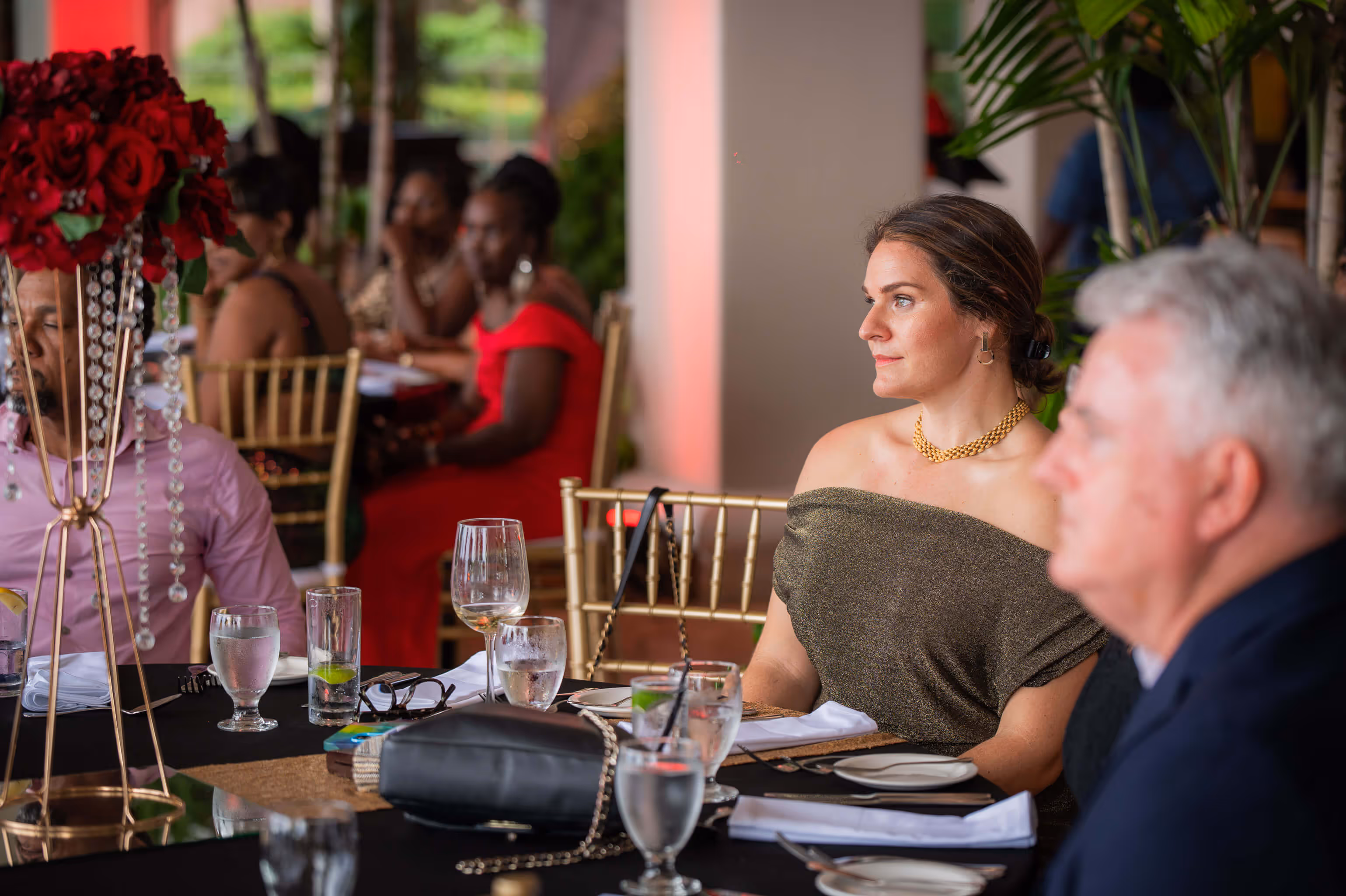 Woman in a shimmery off-shoulder dress and gold necklace sitting at a table during a formal event with other attendees in the background.