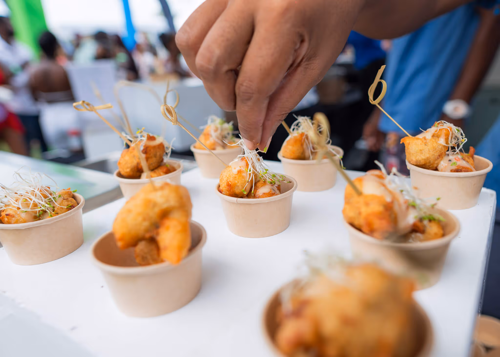 Hand garnishing fried appetizer bites with sprouts in small paper cups on a white tray.