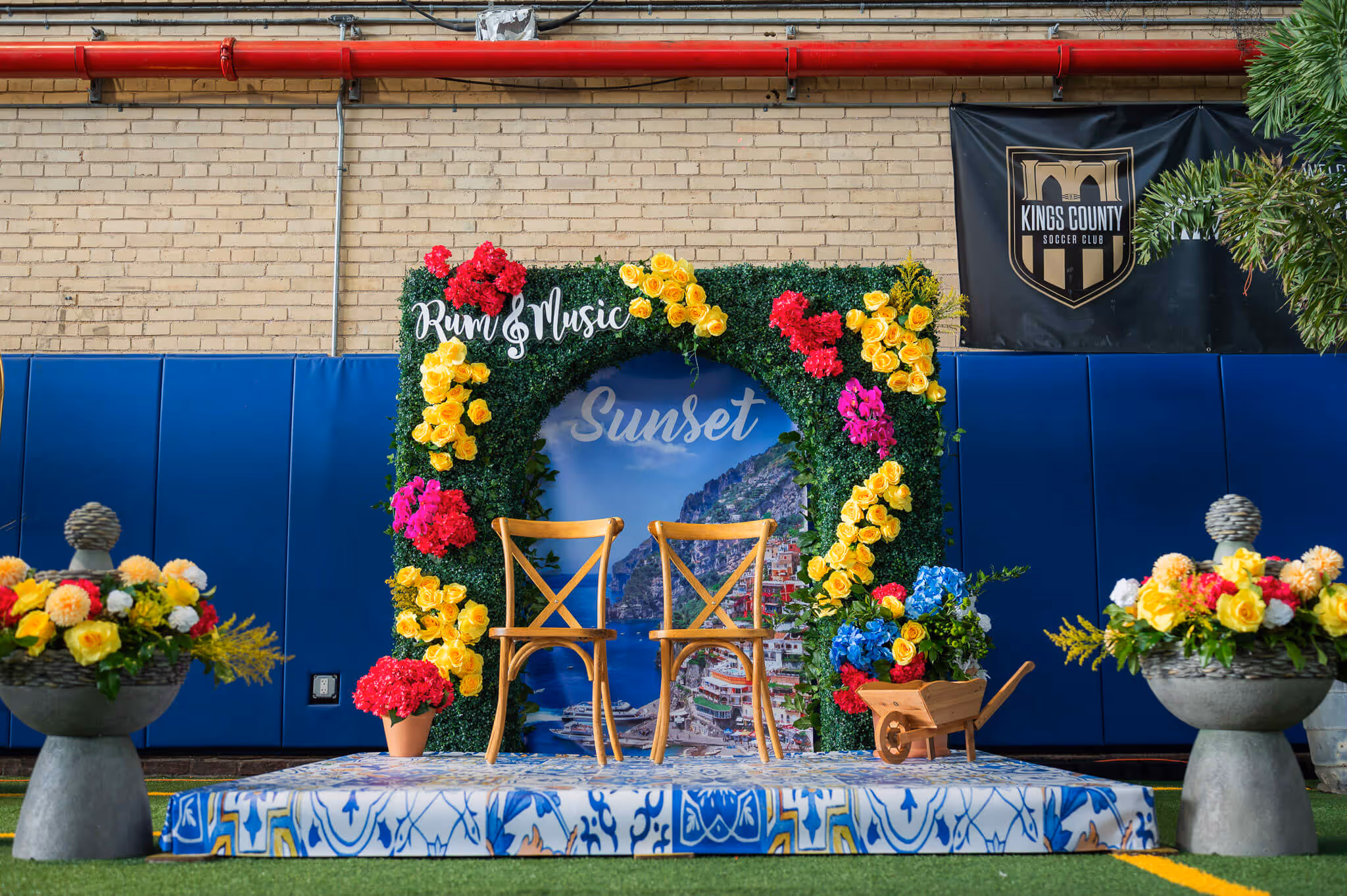 Event setup with two wooden chairs on a decorated blue and white platform under a floral arch reading 'Rum & Music' and 'Sunset', with colorful flowers and a scenic backdrop.