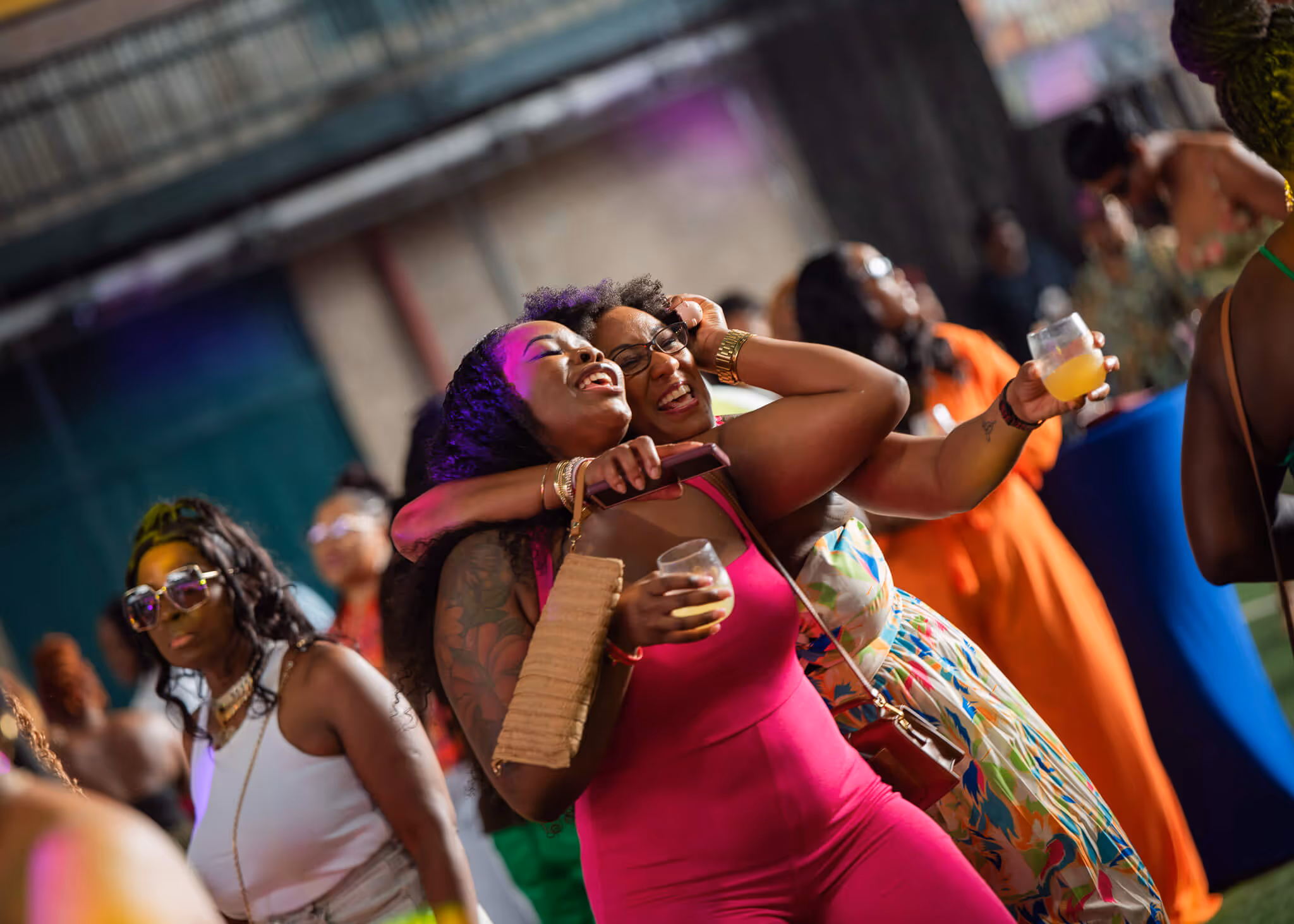 Two women embracing and smiling at a lively party with drinks in their hands.