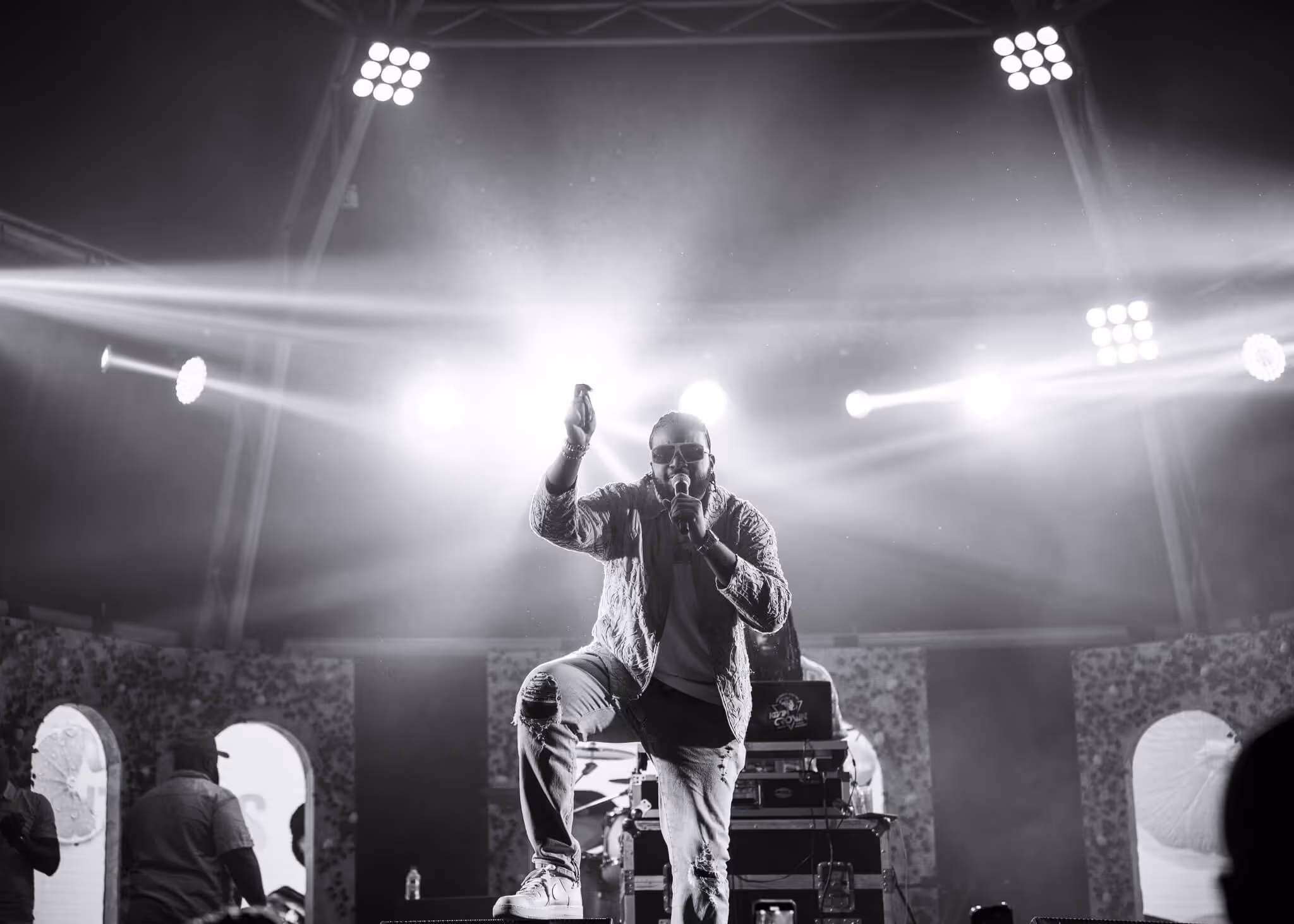 Male performer wearing sunglasses and ripped jeans singing into a microphone on stage with bright spotlights behind him.