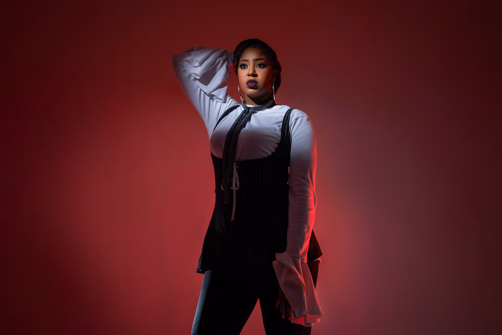 Woman in white blouse and black corset posing with one hand behind her head against a red background.