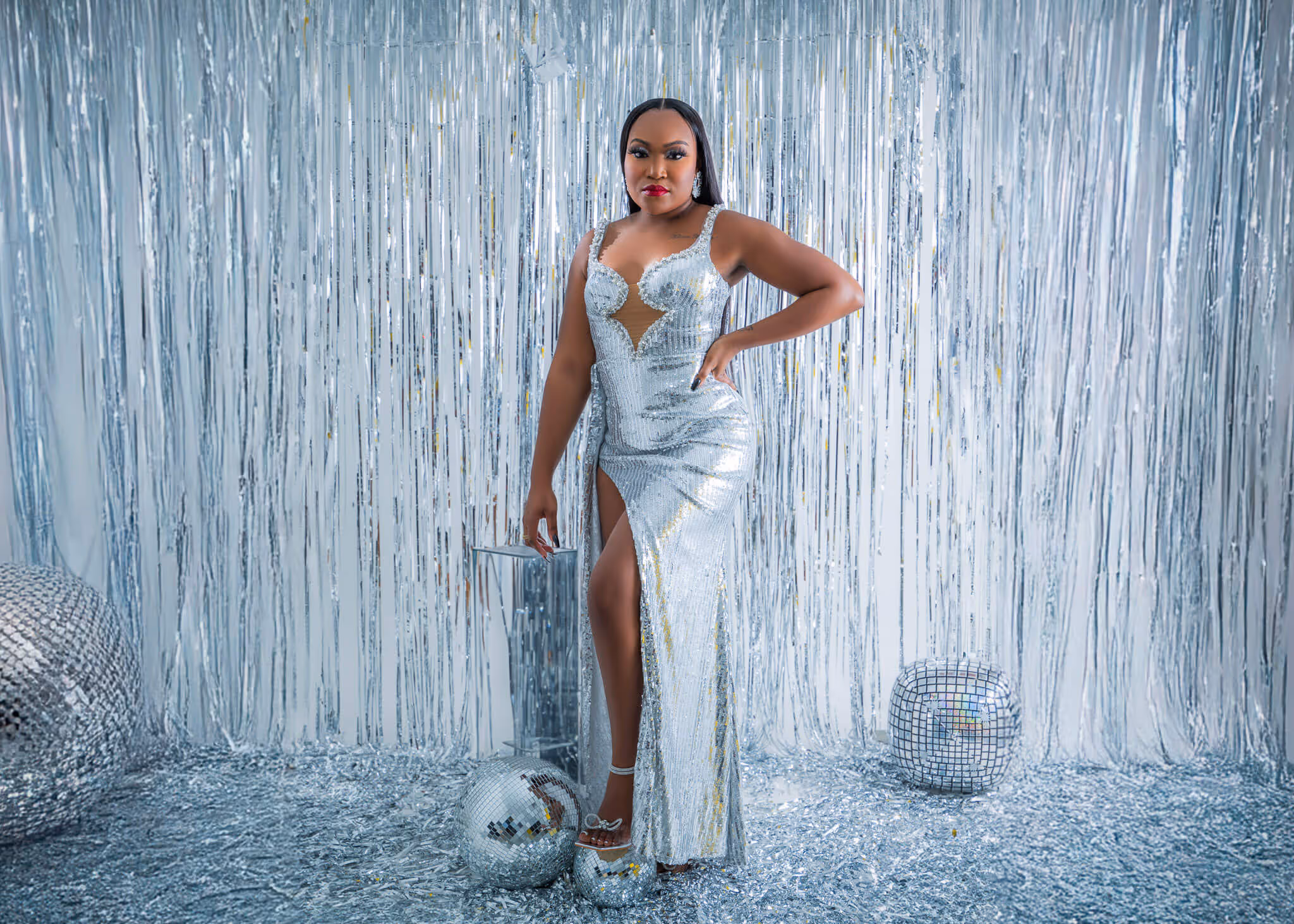 Woman in a silver sequin gown with a high slit standing in a silver tinsel backdrop surrounded by disco balls.