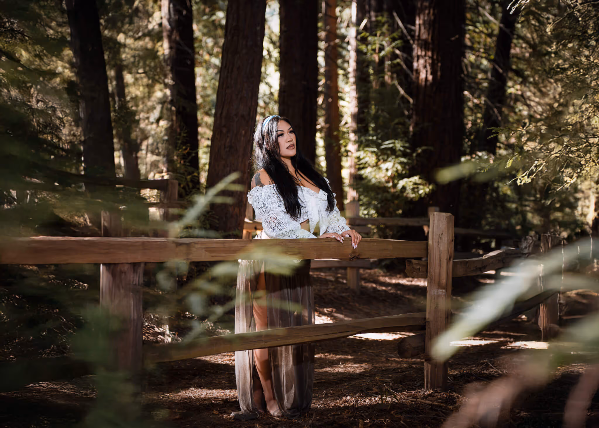 Woman with long dark hair in a white lace top and sheer skirt standing by a wooden fence in a forest.