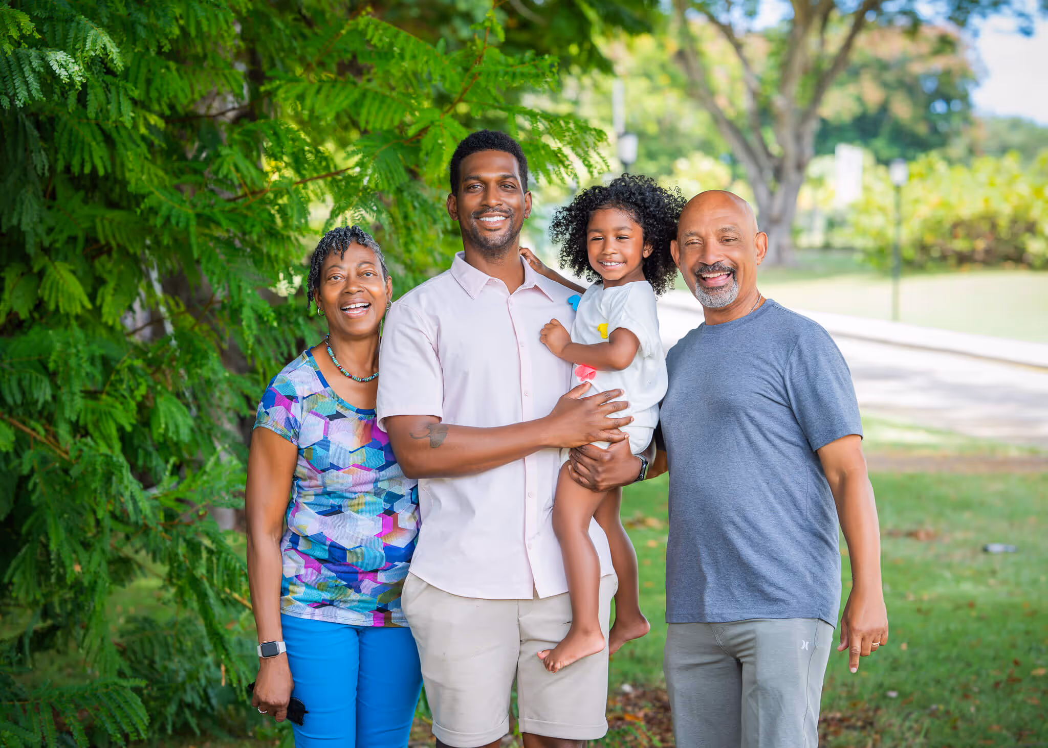 Four smiling family members standing outdoors with green trees in the background.