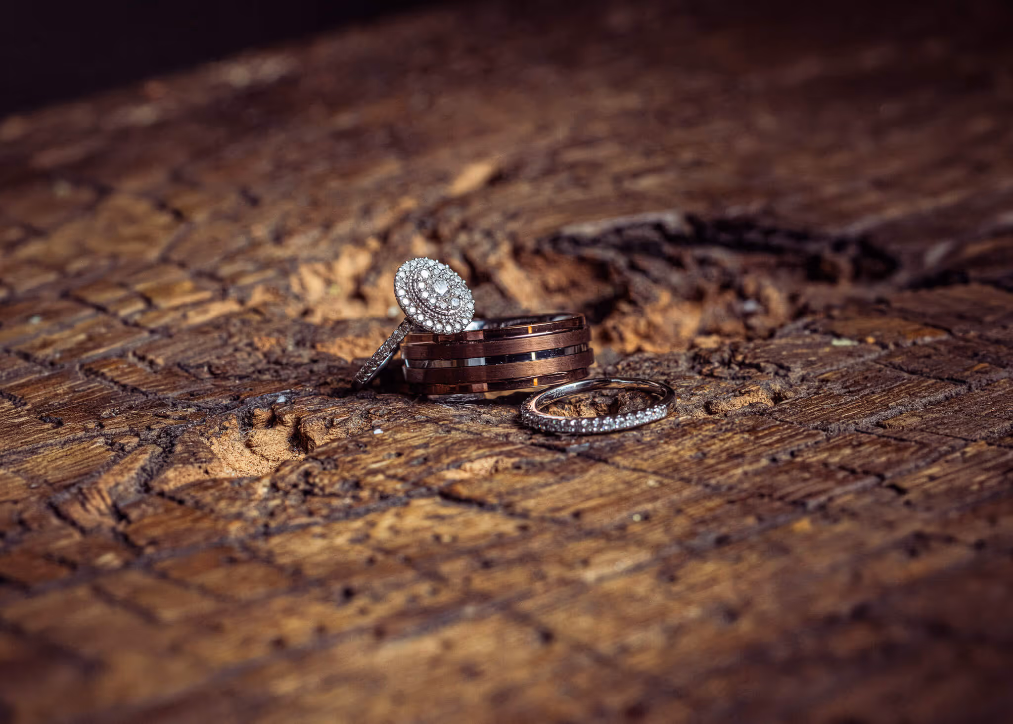 Three rings on rustic wooden surface, including a diamond engagement ring, a wedding band, and a men’s ring with dark inlay.
