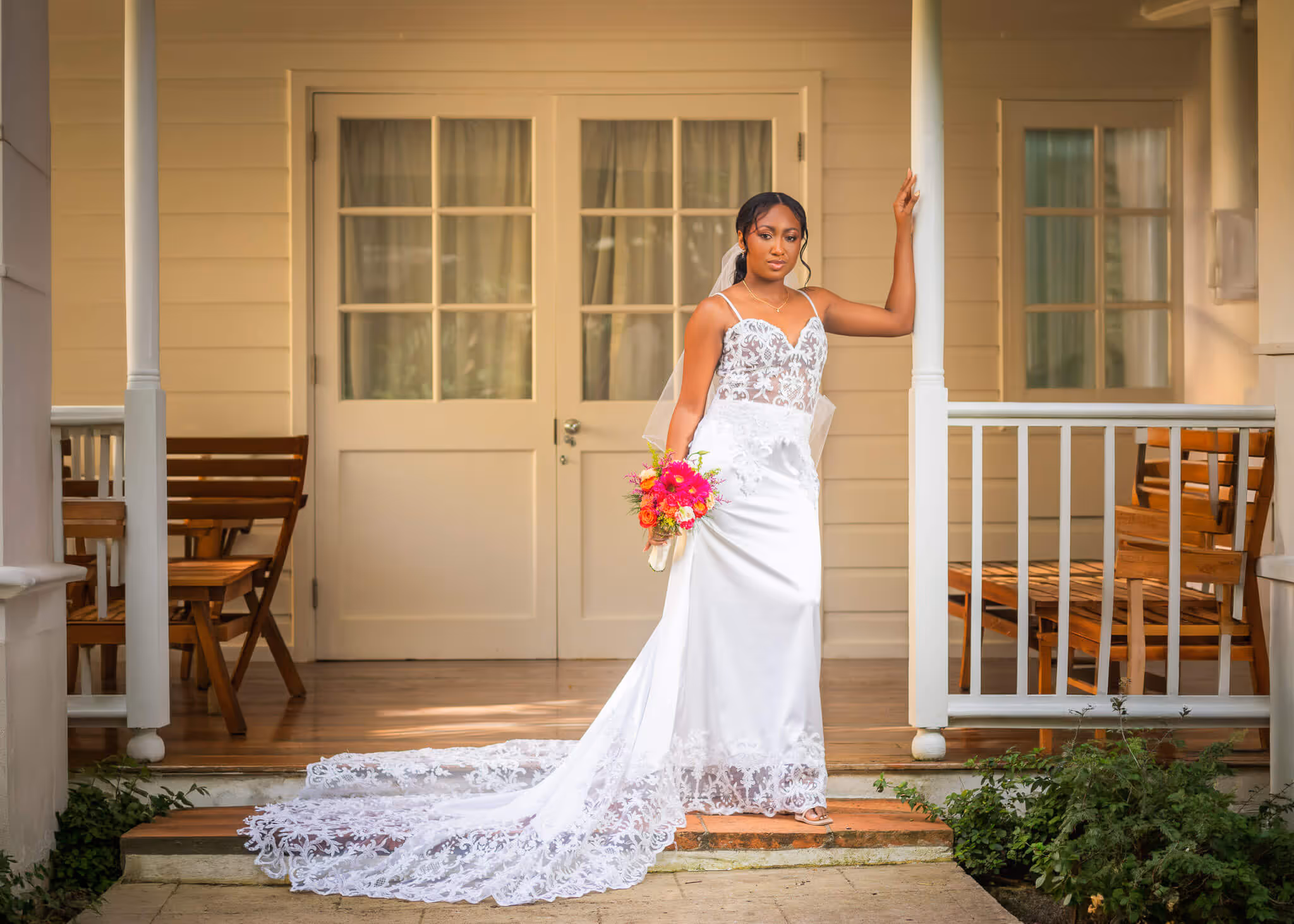 Bride in a white lace wedding dress holding a colorful bouquet, standing on a porch with wooden chairs and a white door in the background.