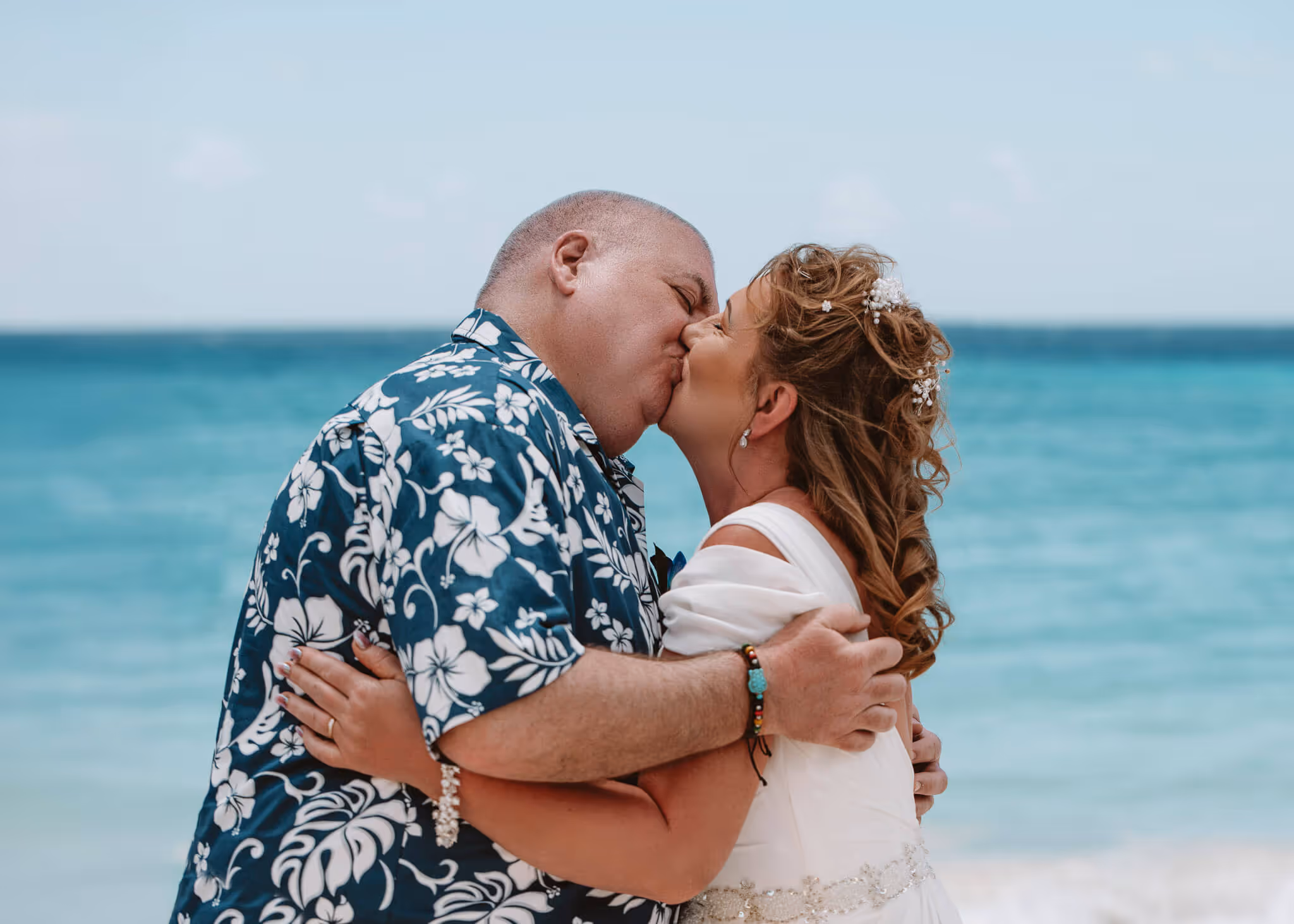 Couple kissing on a beach with the ocean in the background, the woman in a white dress and the man in a blue floral shirt.